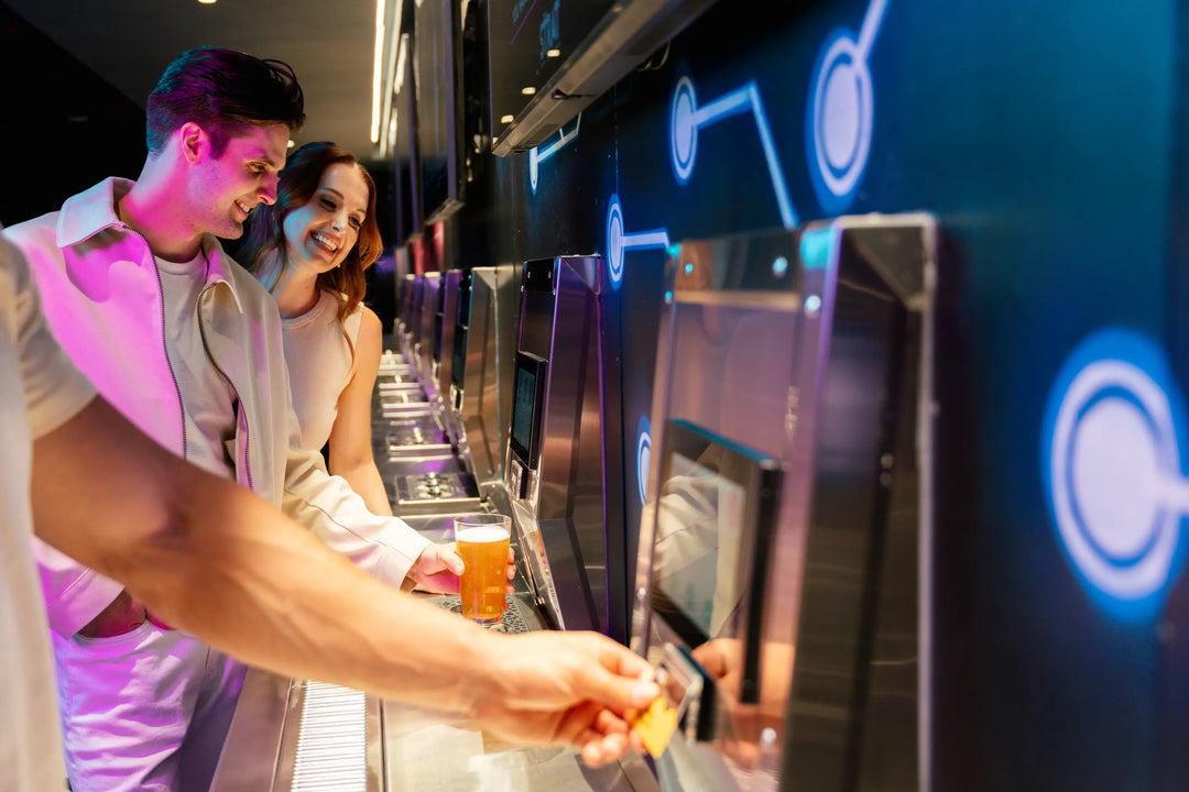 A man and a woman stand at a beverage station with multiple touchscreen drink dispensers. The scene is lit by blue and pink ambient lighting, with one person inserting a card into a dispenser in the foreground.