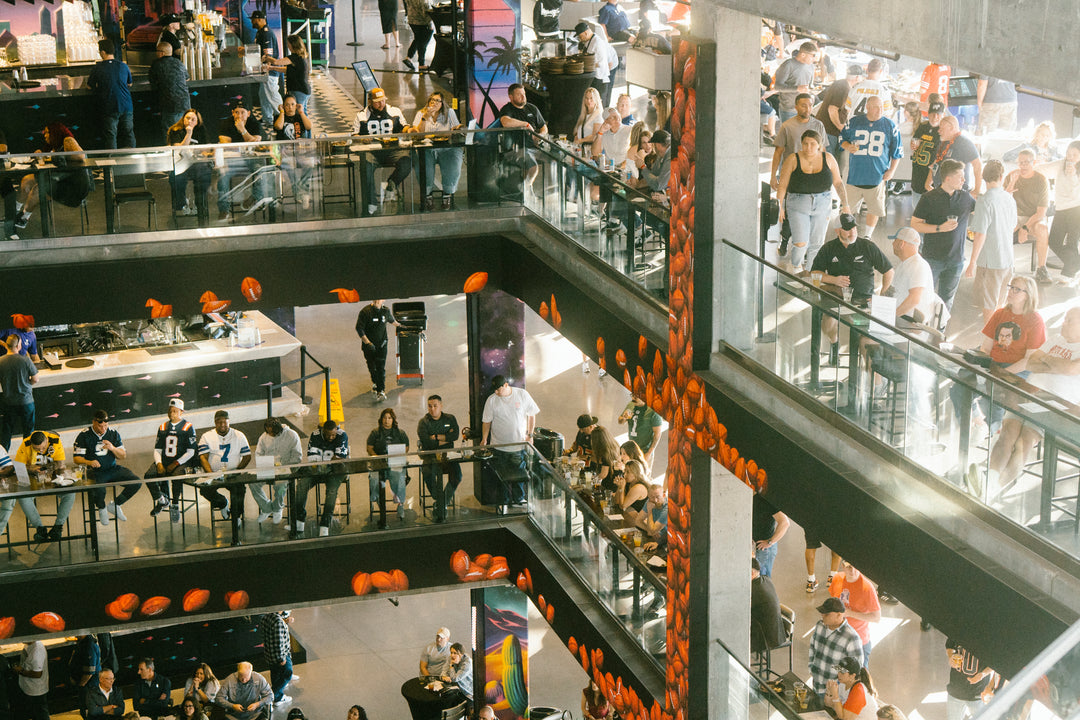 A high-angle view of a crowded multi-level indoor event space at Atomic Golf Las Vegas  with many people walking and standing on balconies, decorated with numerous orange pom-pom balls hanging vertically and along the railings.