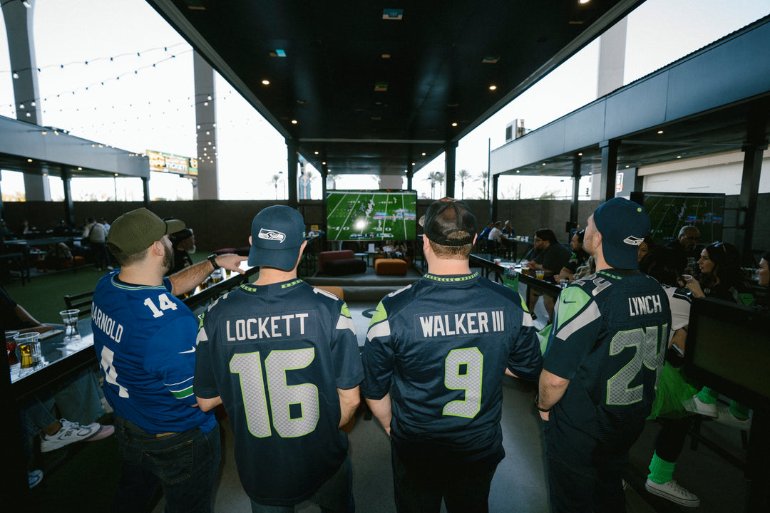 Four men standing in a bar or sports lounge at Atomic Golf Las Vegas, viewed from behind, wearing Seattle Seahawks jerseys with player names and numbers: Lockett 16, Walker III 9, and Lynch 25. A football game is visible on the screen in the background.
