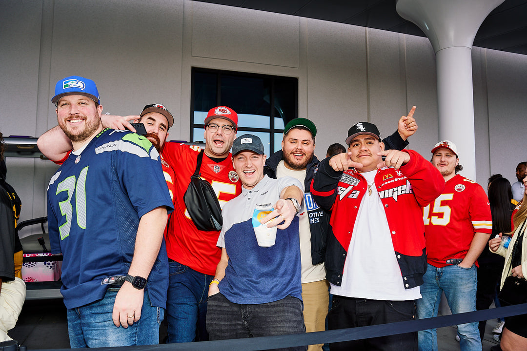 A group of eight sports fans posing together in front of a modern building entrance of Atomic Golf Las Vegas. They are wearing various team jerseys, including blue, red, and white, along with matching caps.
